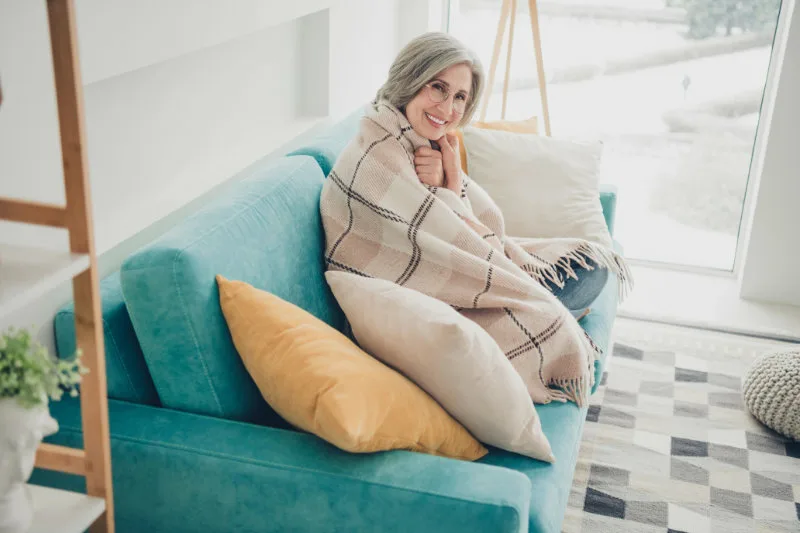 Ältere Frau sitzt eingekuschelt auf dem Sofa - ein Bild für Selbstakzeptanz und Körperfreundlichkeit.